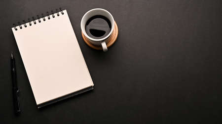 Minimal black workspace table with a cup of coffee, an empty spiral notepad and copy space on black...