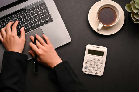 Businesswoman or female accountant working at her office desk, typing on notebook laptop computer. Laptop, calculator and coffee cup on black background. top viewの写真素材