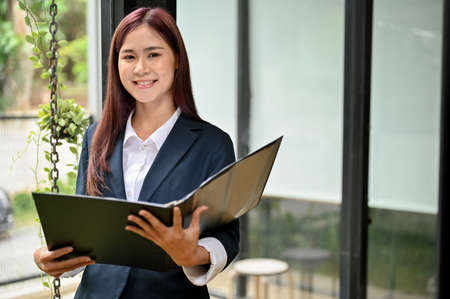 Portrait of a beautiful young asian businesswoman in formal suit with a document folder standing in her modern office.の写真素材