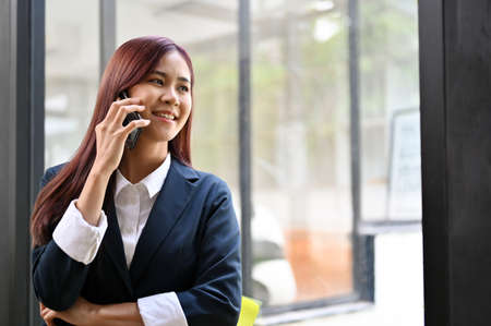 Successful asian businesswoman standing near the office window and talking on the phone with her business supplier.の写真素材