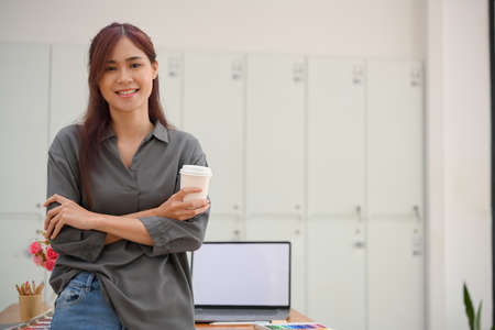 Portrait of a beautiful young asian female graphic designer leaning on her office worktable, crossed arms and holding a coffee cup.の写真素材