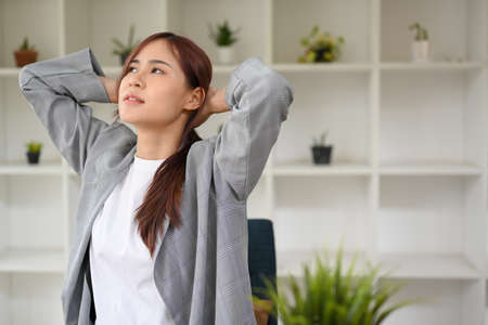 Tired young asian businesswoman or female office worker hand behind her head, stretching after work in front of her office desk.の写真素材