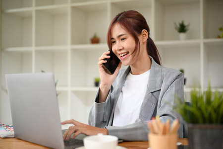 Attractive asian young businesswoman in casual suit talking on the phone with business partner while working at her office desk.の写真素材