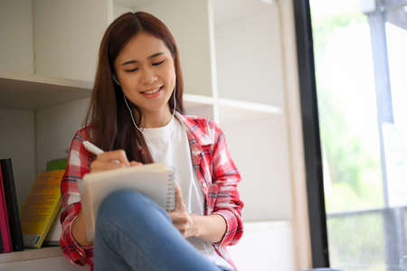 Relaxed young Asian female college student wearing an earphones, listening to music and writing...