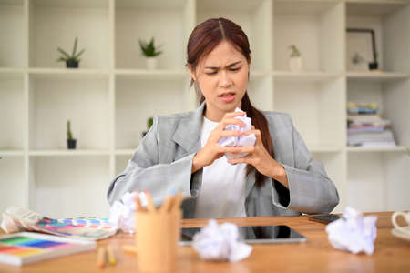 Stressed angry unsatisfied asian businesswoman sits at office desk with crumpled paper and crumpling the document with her hands.の写真素材
