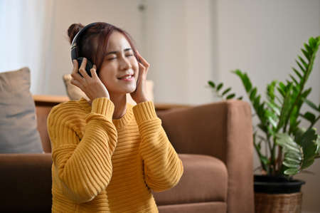 Cheerful millennial Asian female is enjoying the music on her modern wireless headphones in the living room. Leisure and activity conceptの写真素材
