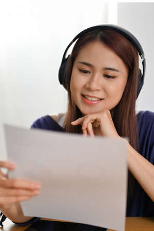 Portrait of a beautiful millennial Asian female with headphones listening her tutor teaching online and reading a content on a paper.の写真素材