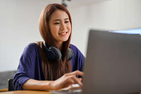 Beautiful and attractive young Asian woman with wireless headphones browsing something on the internet, using portable laptop computer.の写真素材