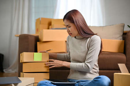 Attractive young asian female online business shop owner packing a customer's orders in her living...