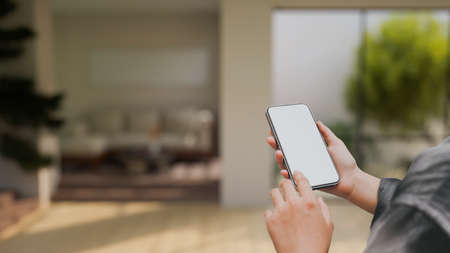 A mockup blank screen of a smartphone in women's hands over blurred modern contemporary living room interior design in the background. 3d rendering, 3d illustrationの写真素材