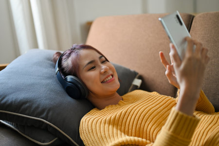 Beautiful millennial asian woman lying on comfortable sofa, listening to music, wearing wireless headphones and using smartphone.の写真素材