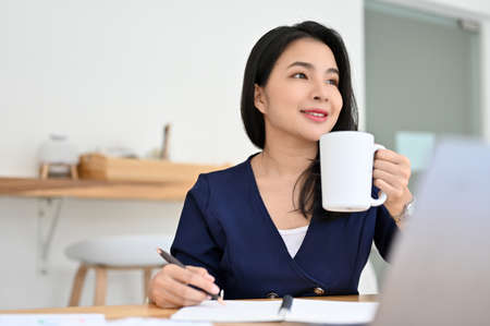Gorgeous and successful young Asian businesswoman or executive manager sits at her office desk sipping morning coffee.の写真素材