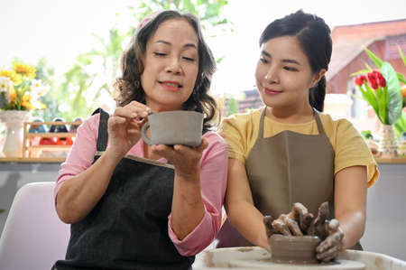 A professional potter teaching and coaching her young female student making a pottery in the workshop. Charming asian woman moulding clay on potter's wheel.の写真素材