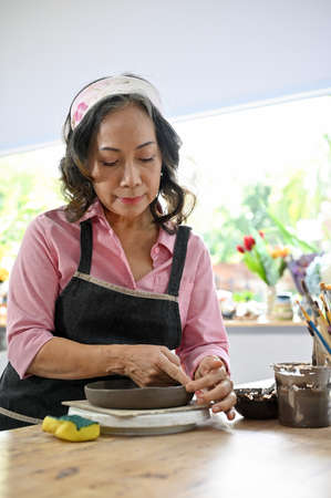Portrait of an asian 60s aged woman with apron making moulding clay ceramic plate in her studio. Handmade ceramic workshop conceptの写真素材