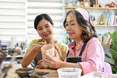 Lovely Asian mom and teens daughter enjoy painting acrylic colour on a ceramic cup together in the handicraft worksop.の写真素材