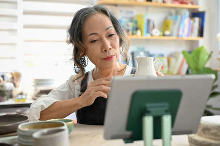 Attractive Asian aged woman looking at tablet screen, holding a ceramic jar, sitting in the workshop...