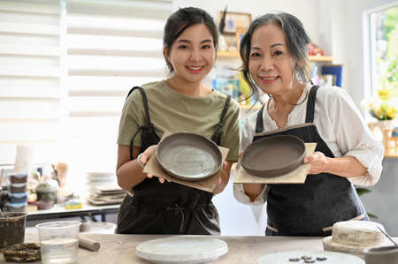 Portrait of a beautiful Asian young female with an attractive asian aged woman holding a handmade bowl and plate in handicraft workshop.の写真素材