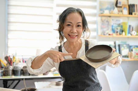 Professional aged Asian female potter showing her ceramic plate in her studio. Beautiful aged Asian woman joining a pottery workshop.の写真素材