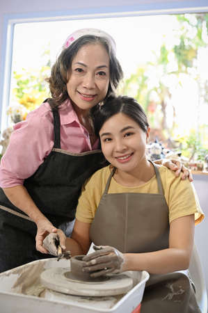 Portrait of a professional aged female craftsman workshop teacher teaching a young female to sculpting pot on a potter's wheel. Handcraft workshop concept.の写真素材