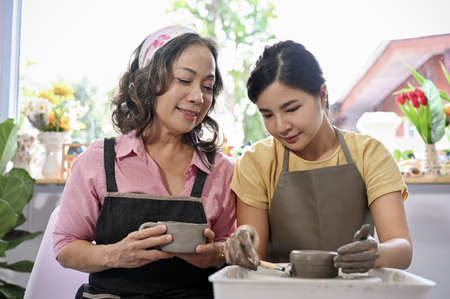 Professional female potter helping and coaching a young female to make a ceramic cup on potter's wheel in the handcraft workshop.の写真素材