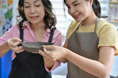 Asian aged woman and young woman focused on molding a handmade ceramic plate together in the...