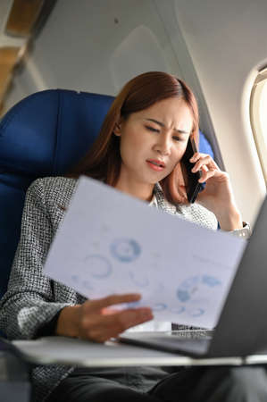 Stressed Millennial asian businesswoman talking on the phone with her secretary, checking her...