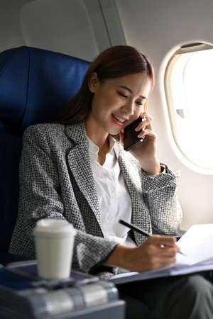 A busy and hardworking Asian businesswoman or female entrepreneur on the phone with a business partner, taking notes on paper while flying to another city for a conference meeting.の写真素材