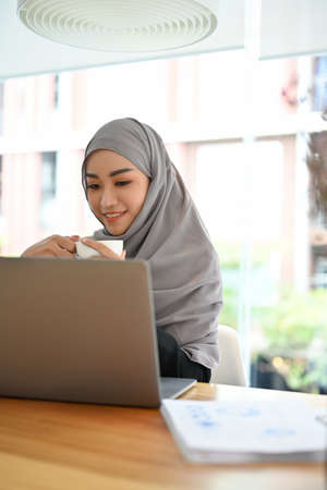 Portrait of a beautiful young asian Muslim businesswoman wearing hijab or head scarf sitting at her office desk with laptop computer.の写真素材