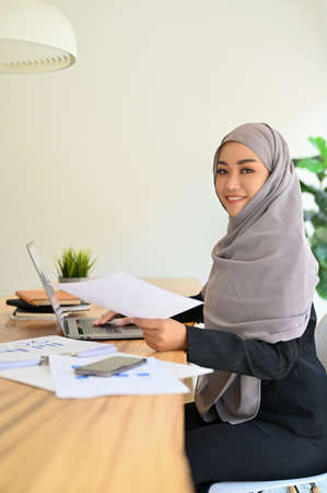 Beautiful and professional Muslim female accountant wearing hijab sits at her office desk holding an accounts paperwork report.の写真素材