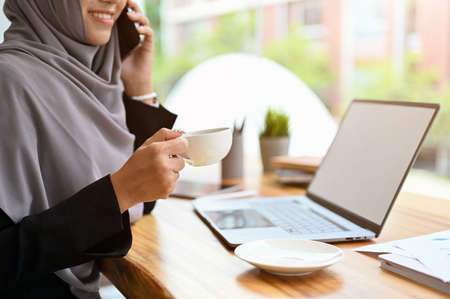 Successful and professional muslim businesswoman wearing hijab on the phone call with business client and sipping a morning coffee at her desk.の写真素材