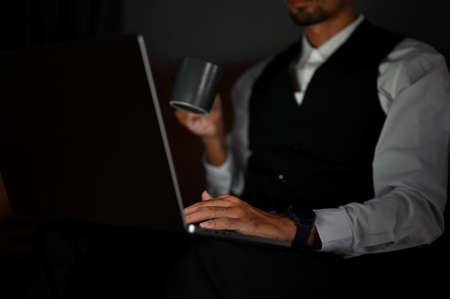 Close-up image and focus hands. A handsome smart male CEO or businessman in formal suit sits on leather sofa sipping coffee and working on his task on laptop computer.の写真素材