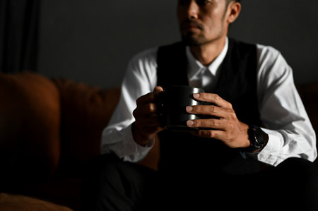 Smart millennial Asian businessman or male executive manager in stylish formal suit sits on dark leather sofa with his coffee. cropped imageの写真素材