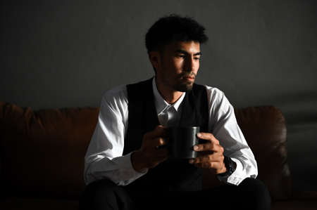 Sad, tired, and bored millennial Asian businessman or male entrepreneur sits on sofa with a cup of coffee, thinking pensively and looking for a work passion.の写真素材