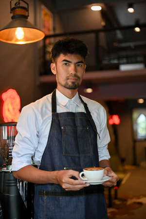 Handsome and professional asian male barista or waitress with apron serving a cup of coffee, standing in the modern coffee shop.の写真素材