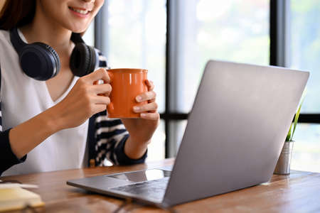 Beautiful young asian woman relaxes sipping a morning coffee while working on her assignment via laptop computer in the coffee shop. Female freelancer remote working in the cafe. cropped imageの写真素材