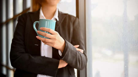Professional Asian businesswoman or female manager in formal suit having a coffee, standing, leaning on the window in the office. cropped imageの写真素材