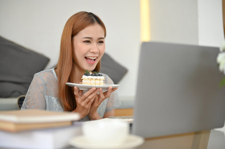 Pretty and cheerful young Asian female food blogger influencer in the cafe reviewing a delicious cake to her online followers via laptop.の写真素材