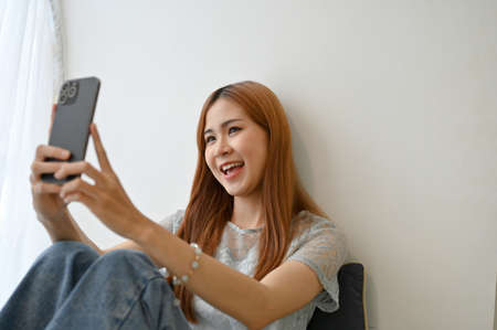 Beautiful millennial Asian girl relaxed in the living room, using her smartphone to take a selfie or...