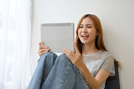 Charming and happy young Asian woman using digital tablet to video call with her friends in the...