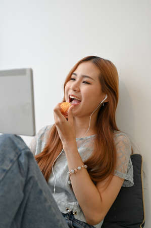 Happy and cheerful millennial Asian girl wearing an earphones enjoy eating cookies while watching something interested on tablet screen.の写真素材