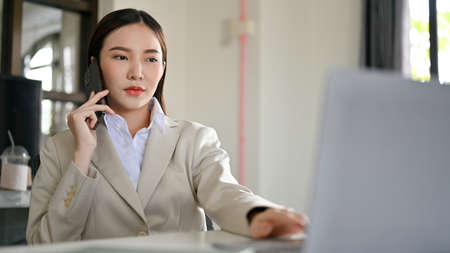 Professional and successful Asian businesswoman or female executive manager in formal suit talking on the phone with her business partner in the office.の写真素材