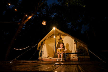 Pretty and relaxed young Asian female camper sitting in front of her tent and reading a book at night. Camping activity concept.の写真素材