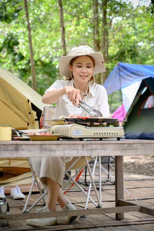 Portrait, Attractive millennial Asian female camper cooking her lunch, roasting a pork chop beef steak on picnic frying pan at the natural campground.の写真素材