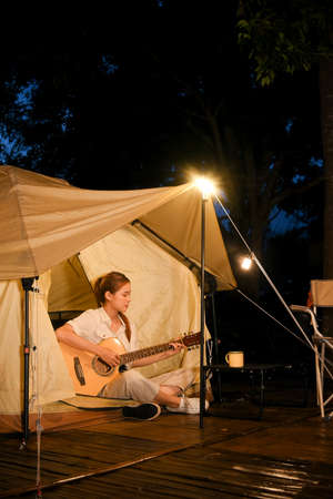 Beautiful young Asian woman singing a song and playing guitar in front of the teepee tent in the...