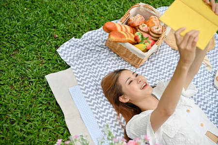 Pretty young Asian female picnicking in the garden, chilling and relaxing on her picnic blanket while reading her favourite book. Top viewの写真素材