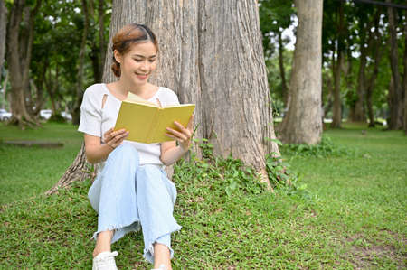 Pretty young Asian female or college student sits under a tree in the park and enjoys reading a book or novel. Outdoor activity conceptの写真素材