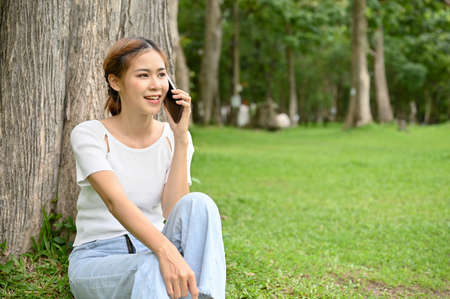 Beautiful young Asian female relaxes while sitting under the tree in the park and on the phone calling someone.の写真素材