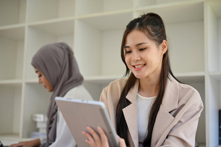 Beautiful young Asian female college student is using a digital tablet to search for some information on the internet while sitting in the campus library co-working space.の写真素材