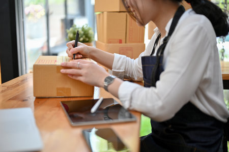 Pretty young Asian female e-commerce startup entrepreneur preparing her shipping packages in the office, writing a customer's address on a cardboard box. cropped imageの写真素材