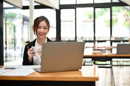 Attractive young Asian businesswoman or female office employee remote working in the coffee shop, having coffee while working her tasks on her portable laptop.の写真素材